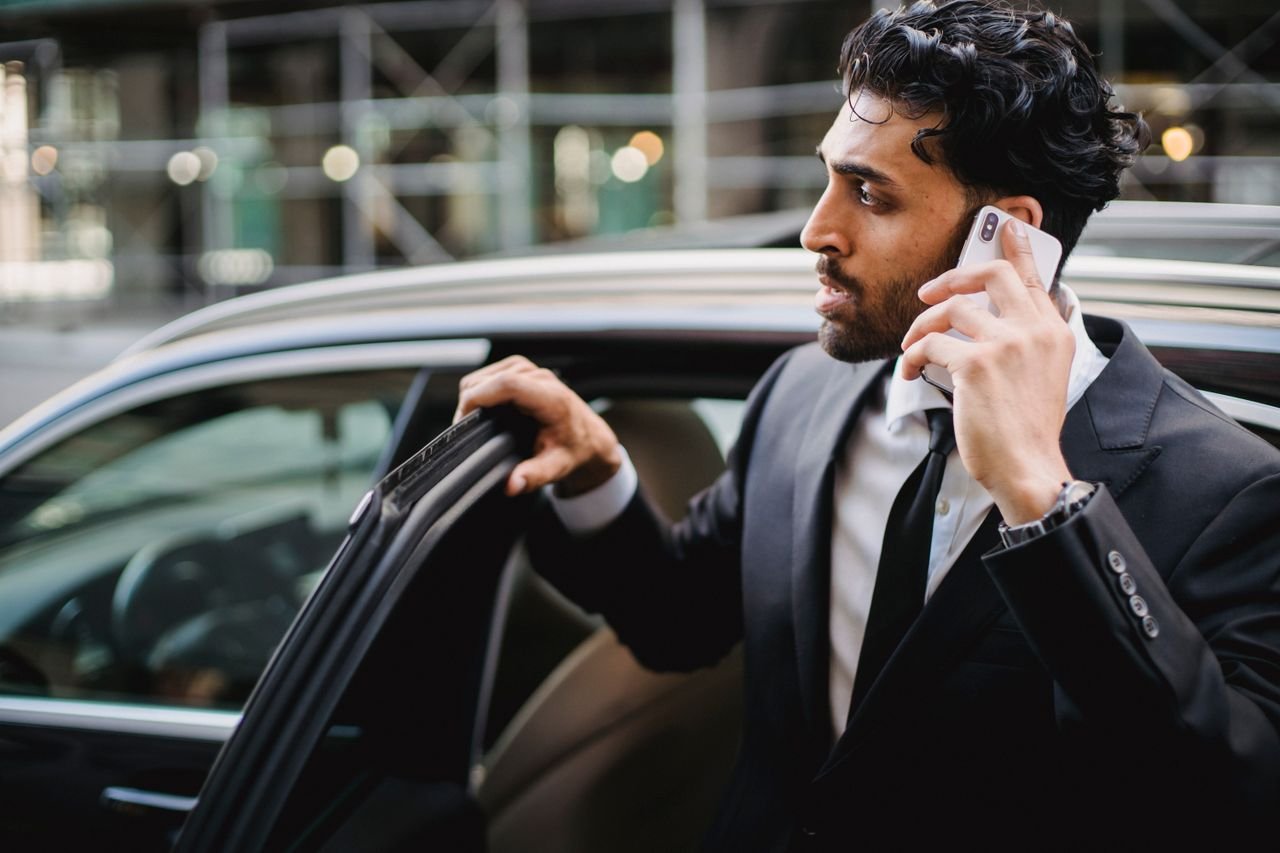 A businessman in a suit talking on his smartphone while exiting a vehicle.